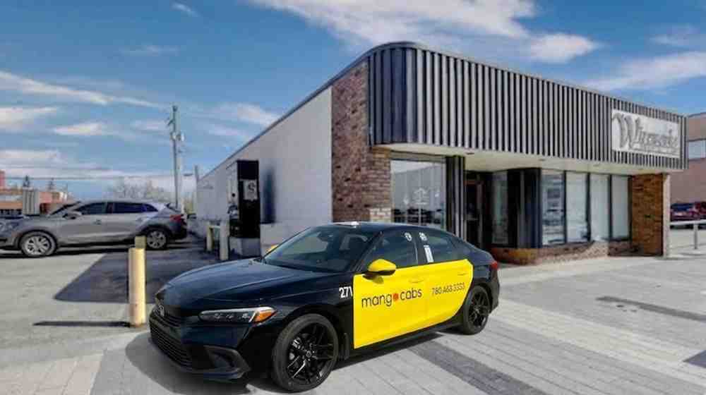 A professional black and yellow Mango Cabs sedan parked on a wide brick sidewalk during the day in front of a modern commercial storefront with a brick and metal facade.